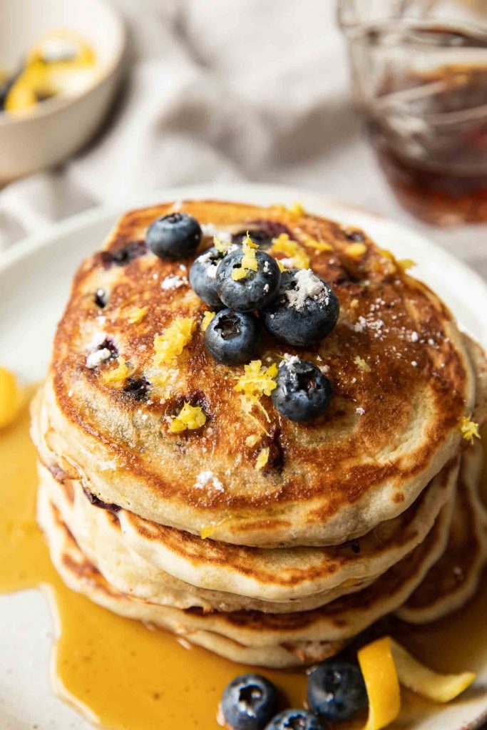 The best lemon blueberry pancakes stacked on a plate with berries, lemon, and powdered sugar.