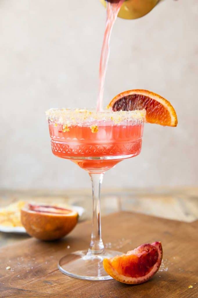 orange margarita pouring into a glass garnished with a blood orange and sugar rim.