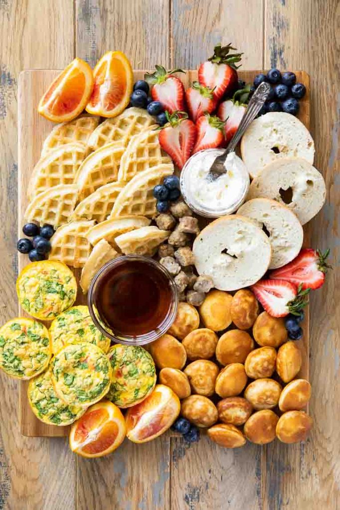Breakfast charcuterie board with fruit, eggs, bagels, waffles, and pancakes.